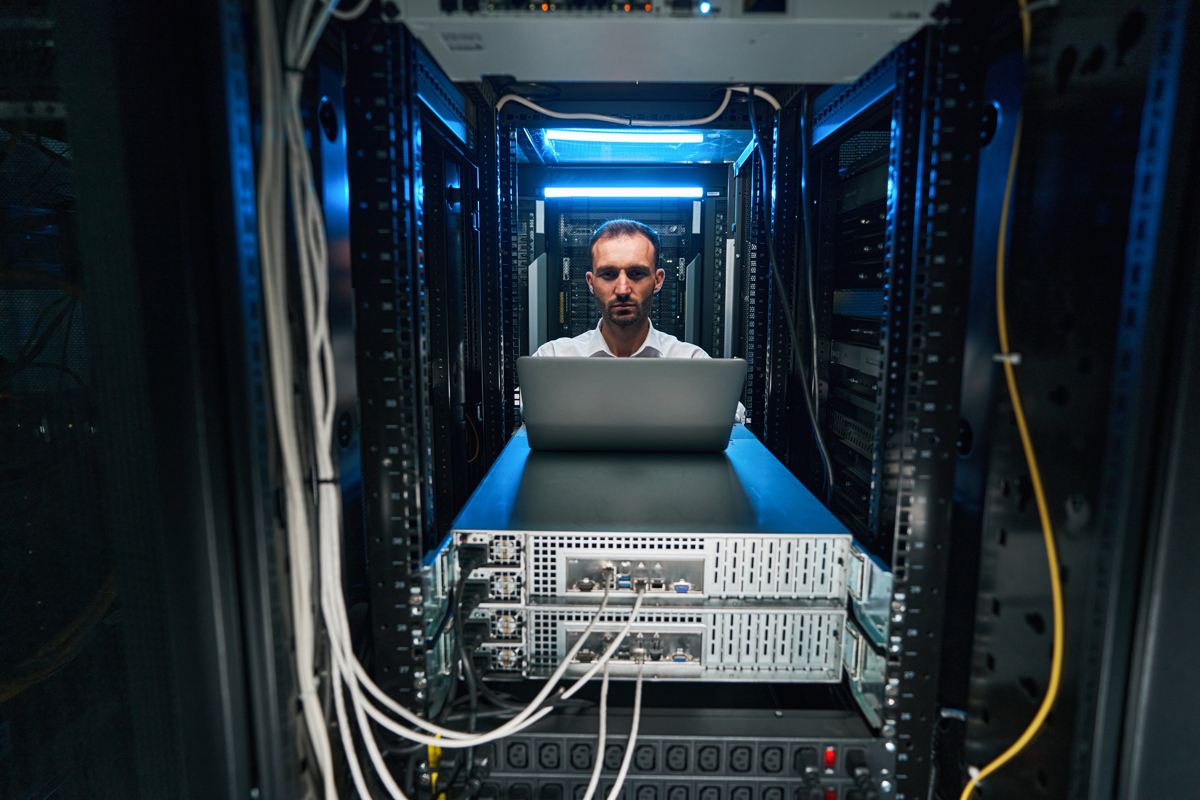 System administrator with laptop in front of him working in server room of data center. (c) AdobeStock