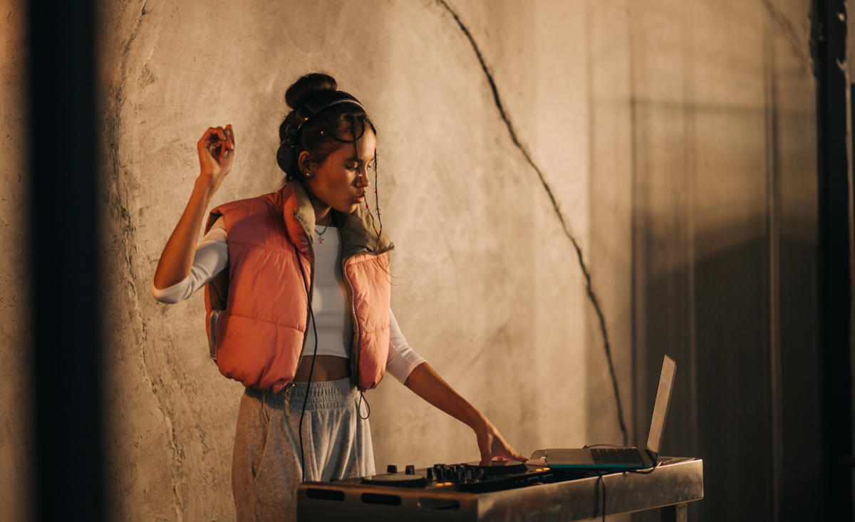 Expressive female DJ in headphones plays music on the remote control and dances on the background of a concrete wall. (c) AdobeStock