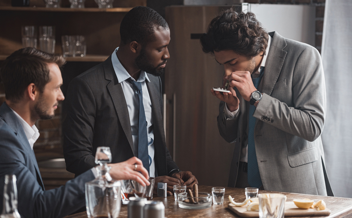 multiethnic men in suits drinking whiskey and looking at friend taking drugs. (c) AdobeStock