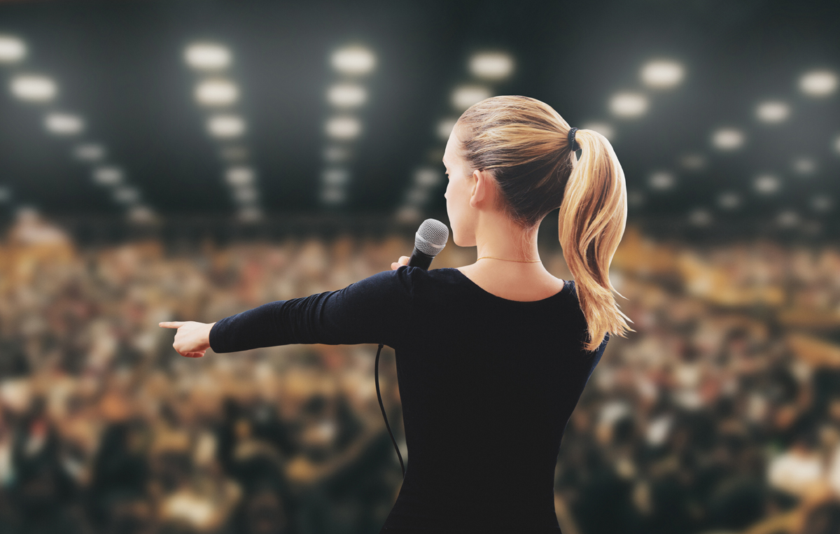 Eine Frau auf einer Bühne in einer großen Halle. (c) AdobeStock