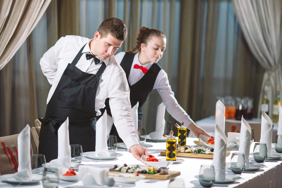 Waiters serving table in the restaurant preparing to receive guests. (c) AdobeStock