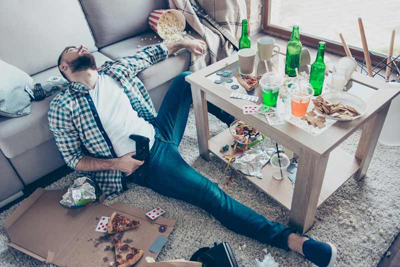 Ein Mann schläft nach einer Party auf dem Boden an eine Couch gelehnt, in der Hand einen Controller, um ihn herum "Partyreste", Stichwort Introducer.
(c) AdobeStock