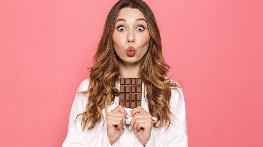 Portrait of a lovely young woman holding chocolate bar isolated over pink background, Stichwort Schokolade.
(c) AdobeStock