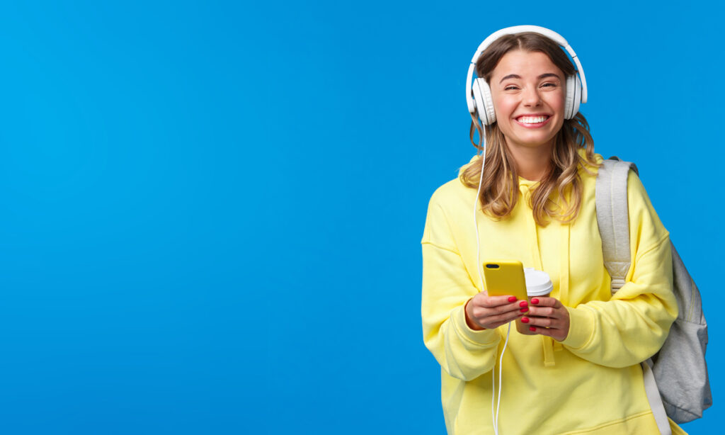 Cheerful pretty young female student with backpack, listening favorite playlist in headphones, laughing and smiling at camera carefree, hold mobile phone, stand blue background, Stichwort lustigste Person.
(c) AdobeStock