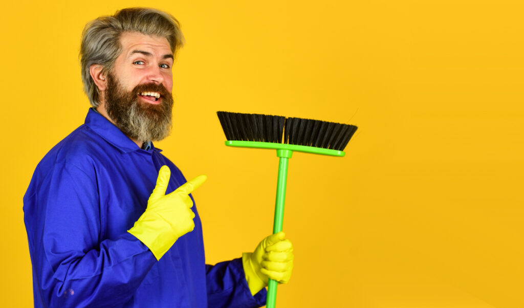 Ein Mann mit gelben Gummihandschuhen zeigt auf einen Besen, den er in der Hand hält, Stichwort Hausarbeit.
(c) AdobeStock