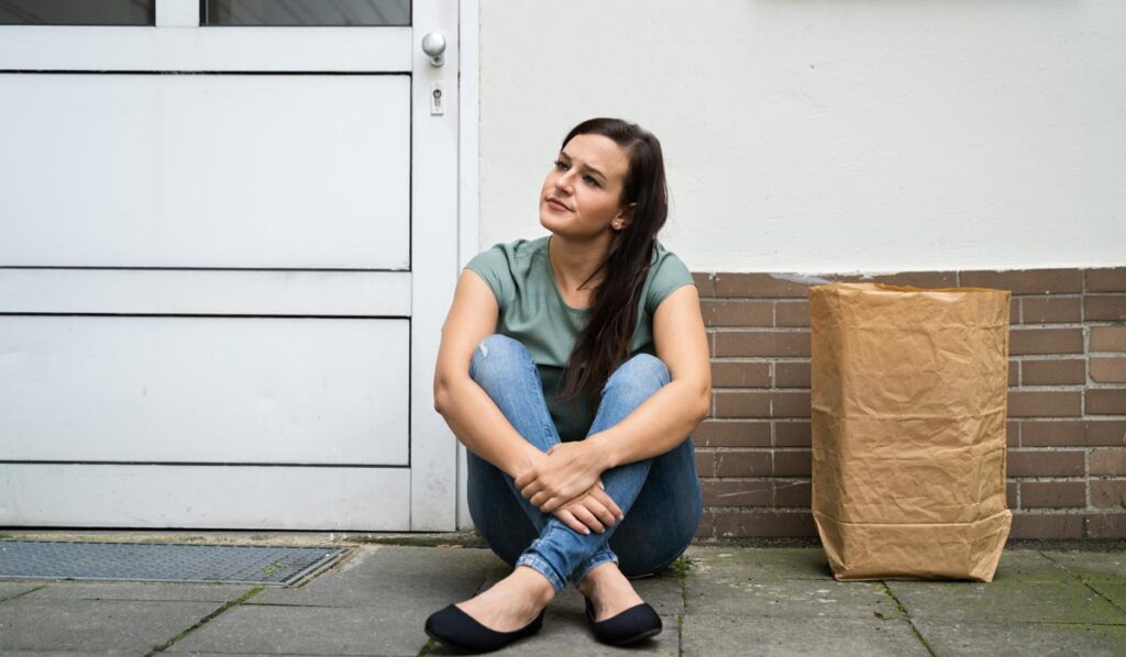 Eine Frau sitzt vor einer Haustür, weil sie den Schlüssel vergessen hat; neben ihr ein großes Einkaufssackerl.
(c) AdobeStock