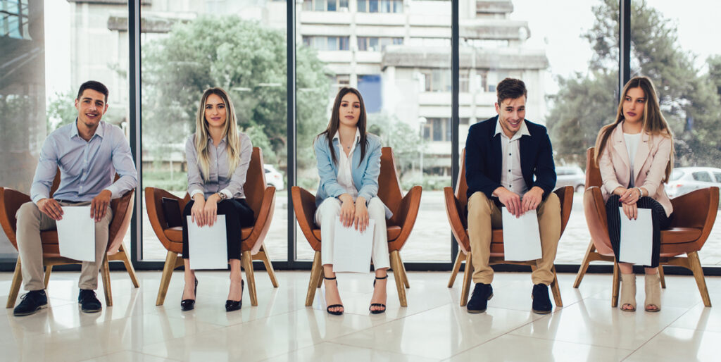 5 Personen sitzen wartend in Sesseln, Stichwort Warteschlangestehen.
(c) AdobeStock