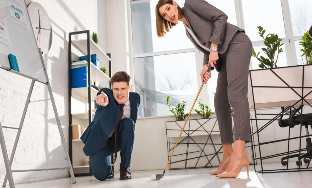 Ein Frau beim Minigolfspielen in einem Büro, neben ihr ein Mann, der auf das Ziel zeigt, Stichwort Prokrastination.
(c) AdobeStock