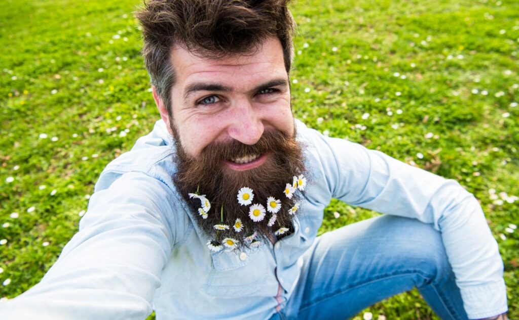 Ein Hippster mit Gänseblümchen im Vollbart sitzt auf einer Wiese und macht ein Selfie.
(c) AdobeStock