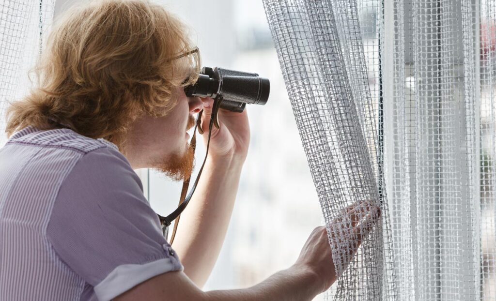 Ein Mann, der mit einem Fernglas aus dem Fenster schaut, Stichwort Spionage.
(c) AdobeStock