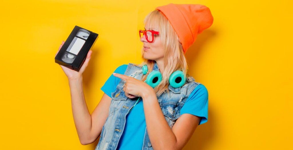 Eine Frau mit roter Brille, oranger Mütze und türkisen Kopfhörern um den Hals zeigt auf eine VHS-Kassette, die sie in der Hand hält, Stichwort Coolsein.
(c) AdobeStock