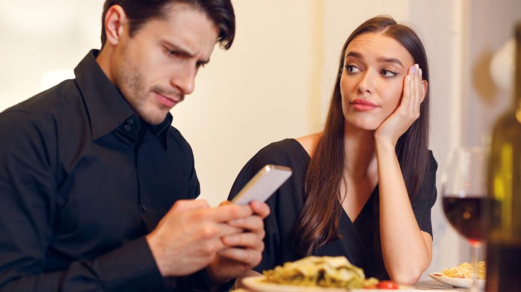 Ein Paar beim Essen. Er tippt am Handy, sie schaut ihn gelangweilt an, Stichwort Paarurlaub.
(c) AdobeStock